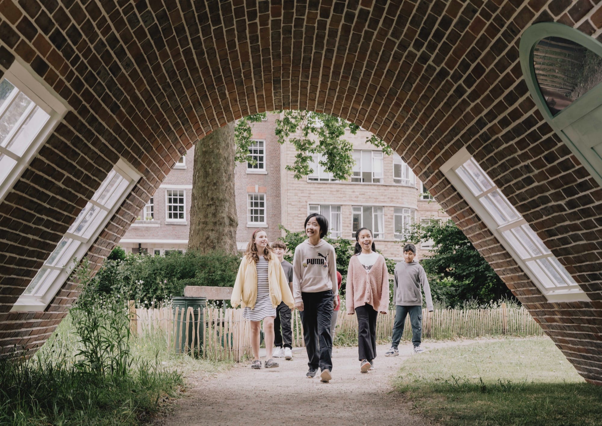 people walk beneath a public artwork resembling a curving brick townhouse facade