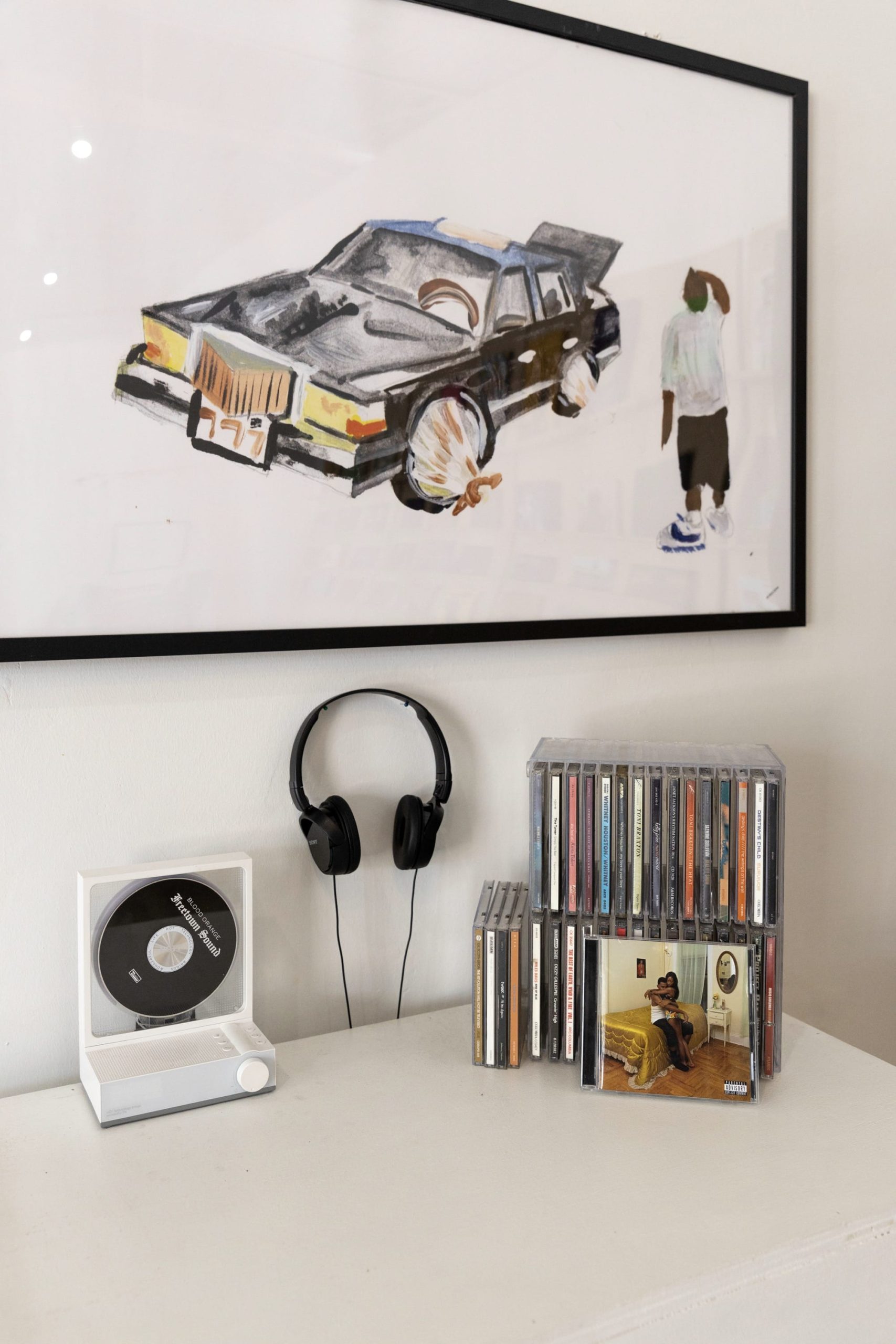 headphones hang on the wall with cds and a player on a white table. an artwork of a man and a car hangs above
