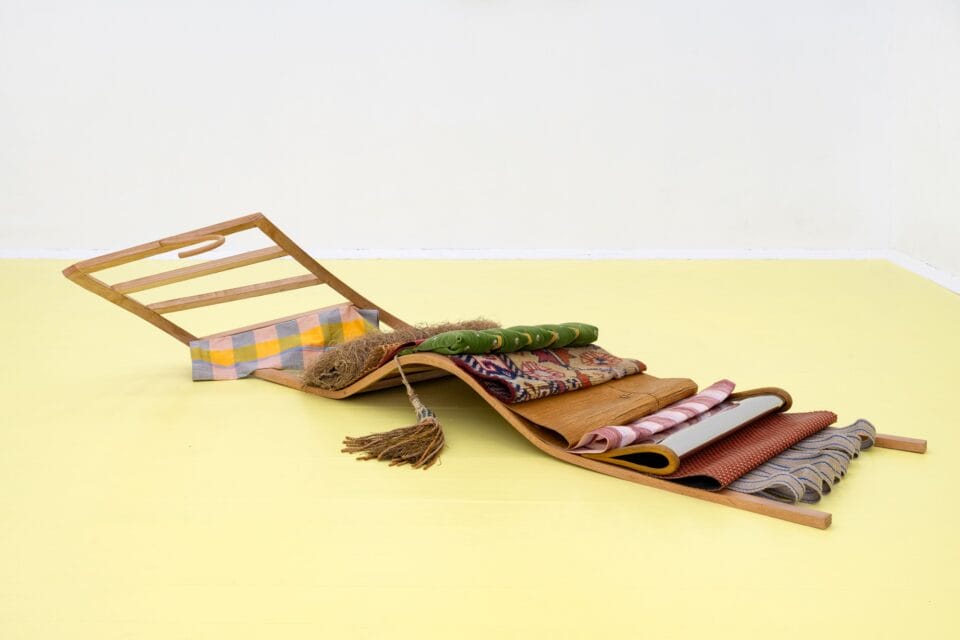 an abstract, mixed-media sculpture of a wooden rack with various textiles and materials draped over it, on the floor